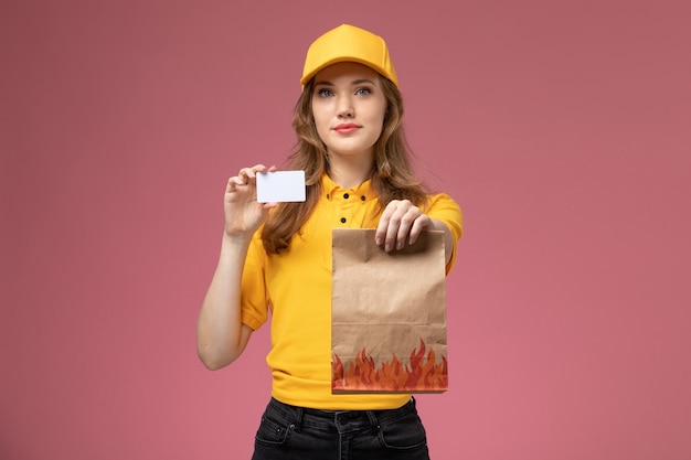 Jovem mensageira de uniforme amarelo segurando um pacote de comida de entrega e um cartão de plástico no fundo rosa.