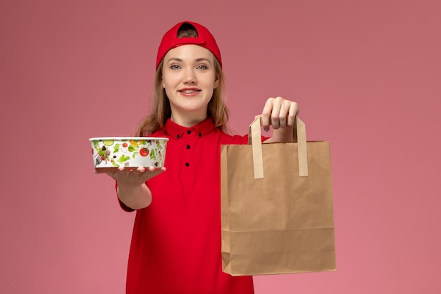 Jovem mensageira de uniforme vermelho, de frente, segurando um pacote de comida para entrega e uma tigela na parede rosa claro