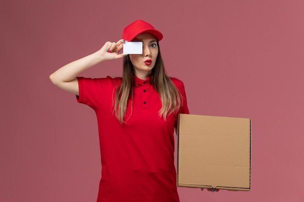 Jovem mensageira de uniforme vermelho segurando uma caixa de comida de entrega e um cartão branco em fundo rosa claro.