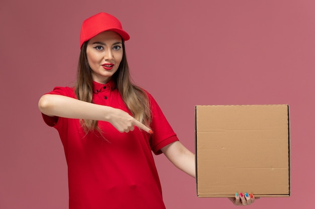 Jovem mensageira de uniforme vermelho segurando uma caixa de comida no fundo rosa.