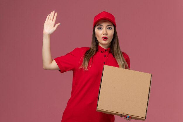 Jovem mensageira de uniforme vermelho segurando uma caixa de entrega de comida na mesa rosa da empresa de entrega de serviço