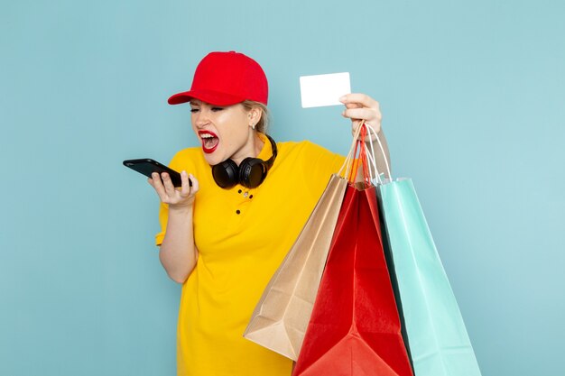 Jovem mensageira feminina de camisa amarela e capa vermelha de frente, segurando pacotes de compras, falando ao telefone sobre o trabalho do espaço azul