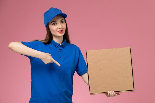 Jovem mensageira feminina de uniforme azul e capa segurando a caixa de comida e abrindo-a na parede rosa