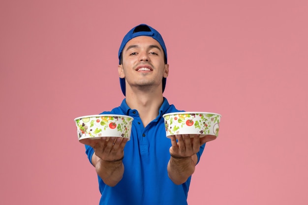 Jovem mensageiro com capa de uniforme azul, frontal, segurando tigelas redondas de entrega na parede rosa claro