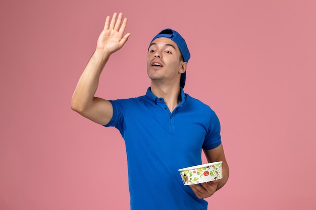 Foto grátis jovem mensageiro com capa de uniforme azul, frontal, segurando uma tigela de entrega redonda na parede rosa claro