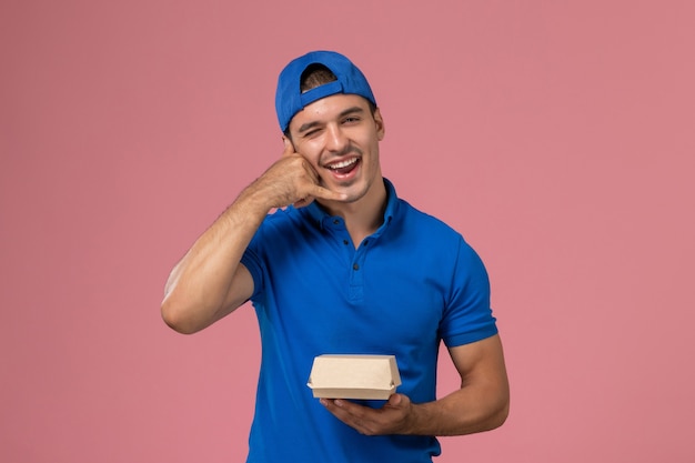 Foto grátis jovem mensageiro com capa de uniforme azul, vista frontal, segurando o pacote de comida de entrega piscando na parede rosa