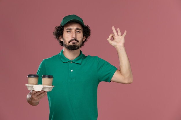 Jovem mensageiro de frente com uniforme verde e capa segurando xícaras de café de entrega no fundo rosa serviço de entrega uniforme de trabalho