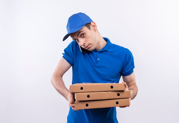 Jovem mensageiro de uniforme azul e boné azul segura caixas de pizza e fala ao telefone