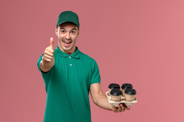 Jovem mensageiro de uniforme verde, de frente, segurando xícaras de café marrons rindo sobre fundo rosa claro