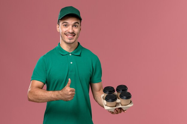 Jovem mensageiro de uniforme verde, de frente, segurando xícaras de café marrons sorrindo sobre fundo rosa claro