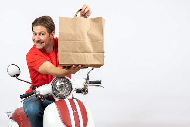 Foto grátis jovem mensageiro feliz em uniforme vermelho sentado na scooter dando uma sacola de papel na parede branca