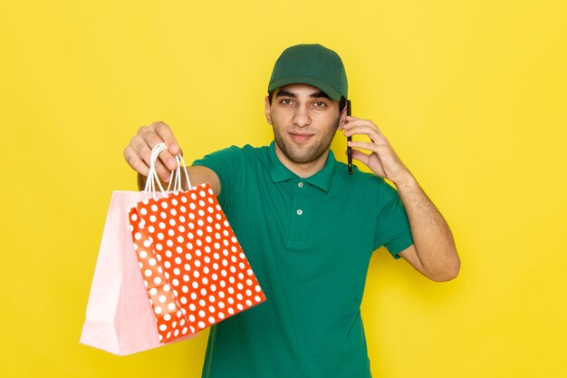 Jovem mensageiro masculino com boné verde de camisa verde falando ao telefone e comprando pacotes no fundo amarelo, entregando a cor do serviço