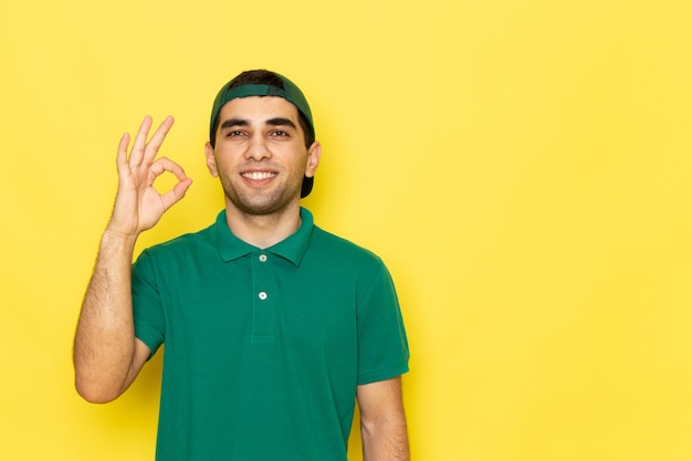 Jovem mensageiro masculino com boné verde de camisa verde sorrindo e mostrando o sinal de tudo bem sorrindo no fundo amarelo.