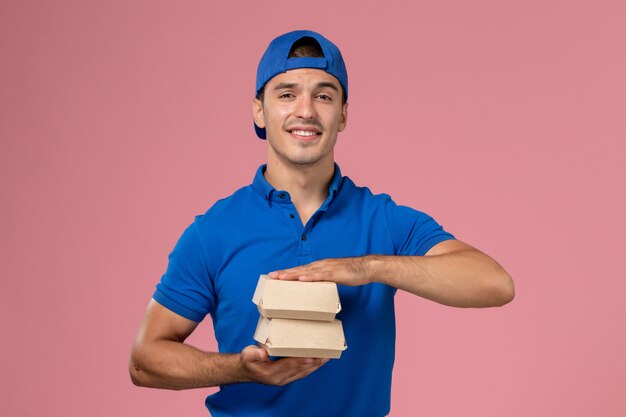 Jovem mensageiro masculino com capa de uniforme azul segurando pequenos pacotes de comida de entrega na parede rosa