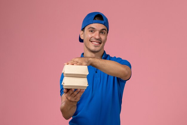Jovem mensageiro masculino com capa de uniforme azul segurando pequenos pacotes de comida de entrega na parede rosa