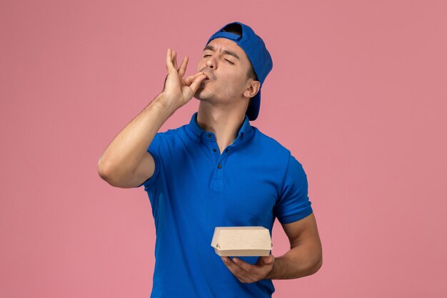 Jovem mensageiro masculino com capa de uniforme azul segurando um pacote de comida de entrega na parede rosa