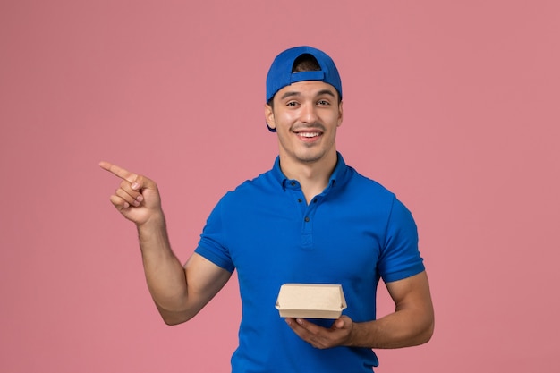 Jovem mensageiro masculino com capa de uniforme azul segurando um pacote de comida de entrega na parede rosa