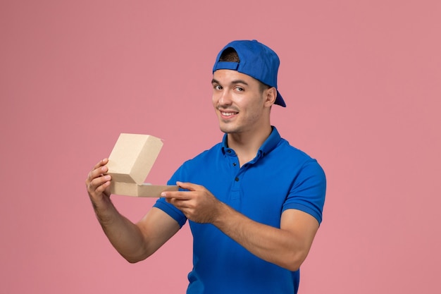 Jovem mensageiro masculino com capa de uniforme azul segurando um pacote vazio de comida na parede rosa