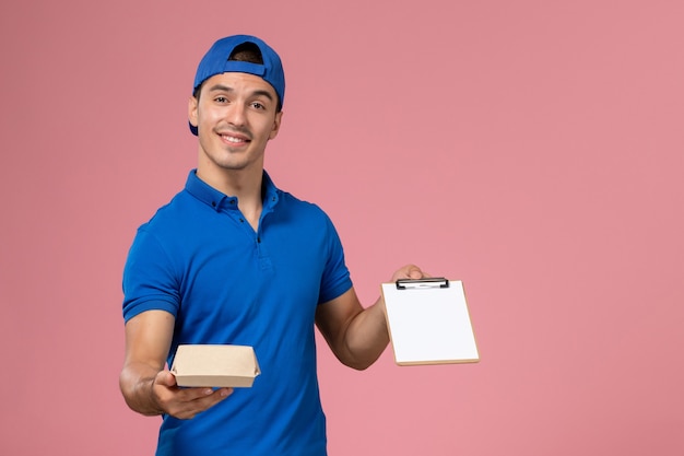 Jovem mensageiro masculino com capa de uniforme azul, vista frontal, segurando um pequeno pacote de comida para entrega e um bloco de notas na parede rosa claro