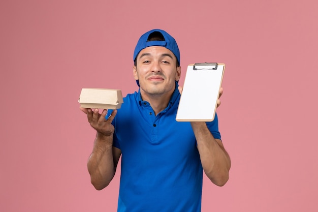 Jovem mensageiro masculino com capa de uniforme azul, vista frontal, segurando um pequeno pacote de comida para entrega e um bloco de notas na parede rosa claro