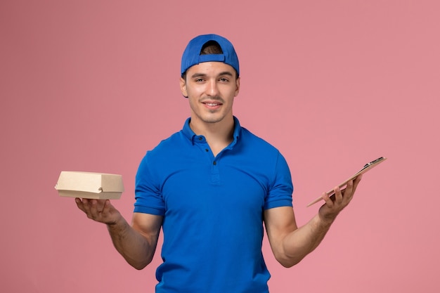 Jovem mensageiro masculino com capa de uniforme azul, vista frontal, segurando um pequeno pacote de comida para entrega e um bloco de notas na parede rosa claro