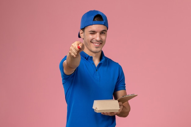 Jovem mensageiro masculino com capa uniforme azul segurando um pacote de comida para entrega e um bloco de notas escrevendo notas na parede rosa claro