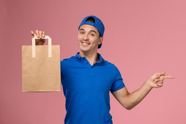 Jovem mensageiro masculino de uniforme azul e capa com pacote de papel de entrega nas mãos na parede rosa