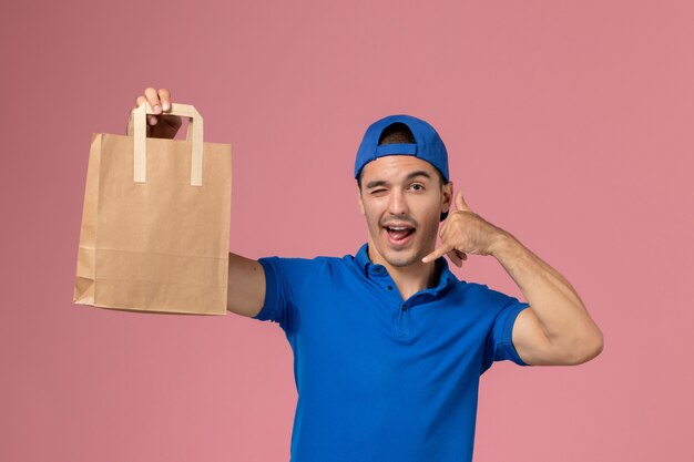 Jovem mensageiro masculino de uniforme azul e capa com pacote de papel de entrega nas mãos na parede rosa