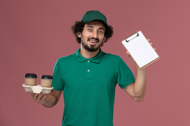 Jovem mensageiro masculino de uniforme verde e capa segurando copos de café de entrega e o bloco de notas no fundo rosa.