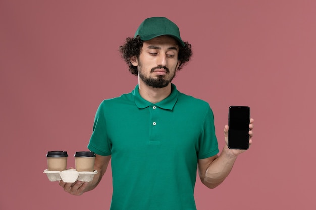 Jovem mensageiro masculino de uniforme verde e capa segurando xícaras de café de entrega e telefone no fundo rosa claro entrega uniforme de serviço