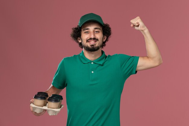 Jovem mensageiro masculino de uniforme verde e capa segurando xícaras de café marrom flexionando fundo rosa claro serviço uniforme entregador