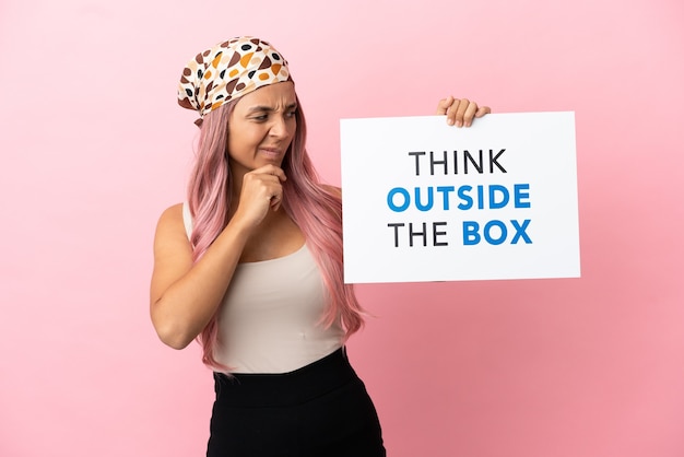 Jovem mestiça com cabelo rosa isolada em um fundo rosa segurando um cartaz com o texto pense fora da caixa e pensando