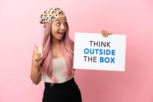 Jovem mestiça com cabelo rosa isolada em um fundo rosa segurando um cartaz com o texto pense fora da caixa e pensando