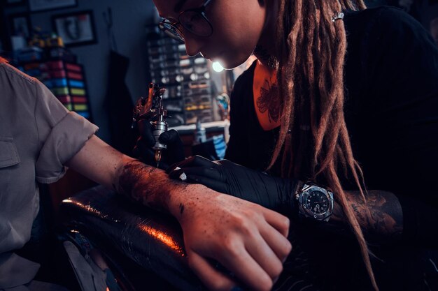 Jovem mestre de tatuagem focado está fazendo tatuagem na mão do cliente no salão de tatuagem escuro.