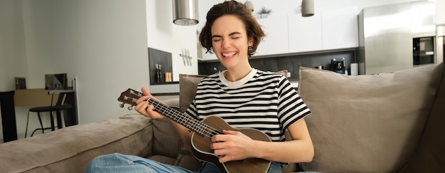 Foto grátis jovem modelo despreocupada tocando ukulele de concerto castanho cantando com paixão sentada no sofá com