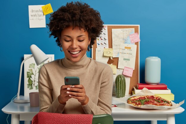 Jovem modelo encantada com cabelo encaracolado usa o celular para bater um papo e navegar na internet, aproveita o tempo livre depois de terminar o trabalho