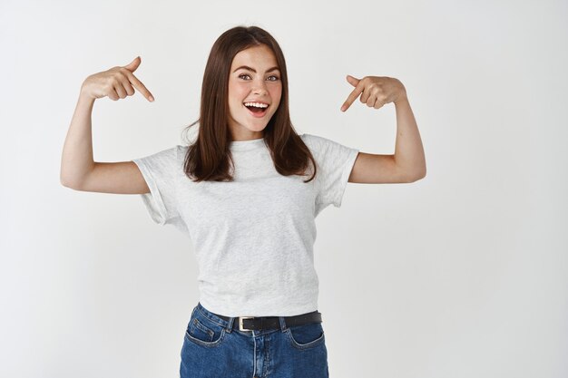 Jovem modelo feminino mostrando o anúncio, apontando o dedo para baixo no logotipo da camiseta e sorrindo, em pé sobre uma parede branca.