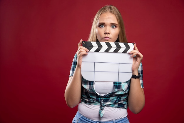 Foto grátis jovem modelo loira segurando um claquete de filmagem de filme em branco e parece estressada e inexperiente.