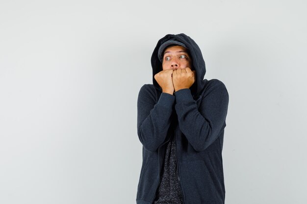 Jovem mordendo os punhos e sentindo frio em t-shirt, jaqueta, boné e parecendo humilde. vista frontal.