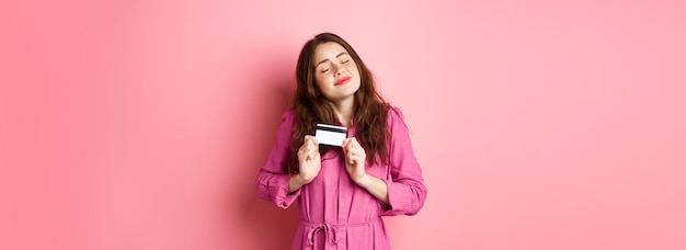 Jovem morena abraçando seu cartão de crédito com prazer sorrindo satisfeito em pé contra as costas rosa
