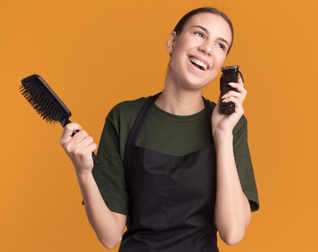 Jovem morena alegre barbeiro de uniforme segurando uma tesoura de cabelo e um pente olhando para o lado