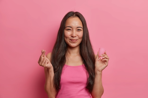 Jovem morena asiática posa com copo menstrual, mostra como usar, dá recomendações, faz sinal coreano, isolado na parede rosa. Saúde da mulher, alternativa zero de resíduos e ginecologia
