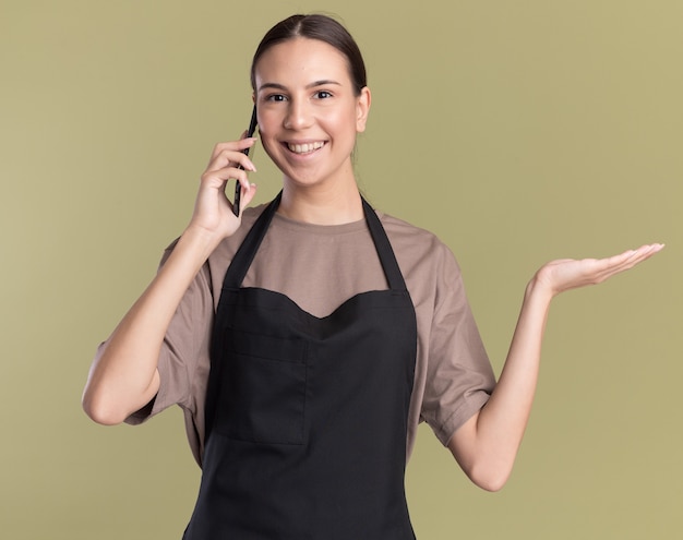 Jovem morena barbeiro de uniforme sorridente com a mão aberta e fala ao telefone