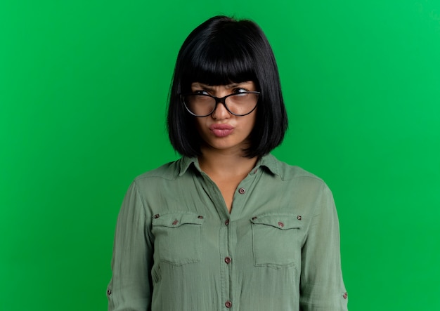 Jovem morena caucasiana séria com óculos ópticos olha para o lado isolado em um fundo verde com espaço de cópia