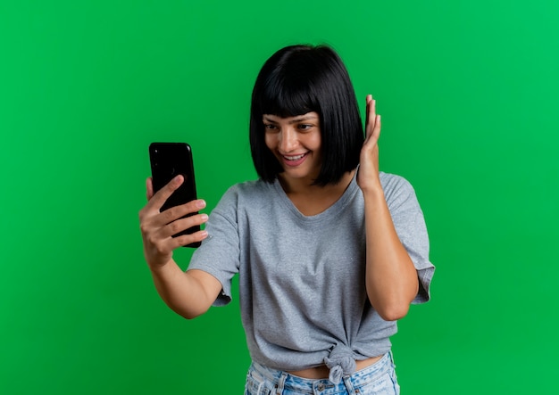 Foto grátis jovem morena caucasiana sorridente levanta a mão e olha para o telefone isolado em um fundo verde com espaço de cópia