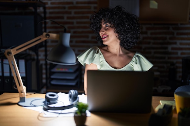 Foto grátis jovem morena com cabelo encaracolado trabalhando no escritório à noite olhando para o lado com sorriso no rosto expressão natural rindo confiante