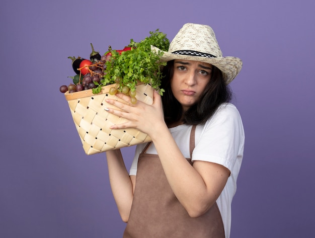 Jovem morena decepcionada, de uniforme, usando chapéu de jardinagem, segurando uma cesta de vegetais isolada na parede roxa