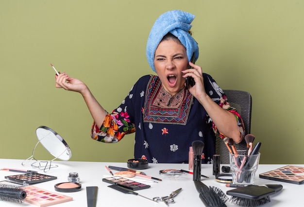 Foto grátis jovem morena irritada com o cabelo enrolado em uma toalha, sentada à mesa com ferramentas de maquiagem, gritando com alguém no telefone e segurando um brilho labial isolado na parede verde oliva com espaço de cópia