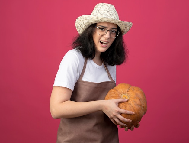 Jovem morena irritada com o uso de óculos e uniforme, usando um chapéu de jardinagem, segurando uma abóbora isolada na parede rosa