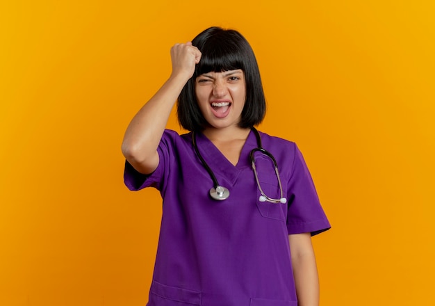 Jovem morena irritada com uma médica de uniforme com estetoscópio coloca o punho na cabeça isolada em um fundo laranja com espaço de cópia